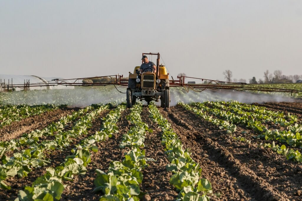 A farmer on a tractor sprays crops with fertilizer in a rural agricultural setting.